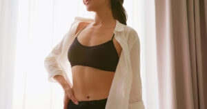 Woman in a black sports bra and open white shirt showing her abdomen, illustrating areas addressed by mini and full tummy tuck surgery.